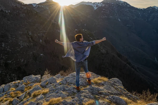 Happy motivated and inspired man with spread hands stands on the edge of rocky cliff watching sunset in the mountains. Motivation and inspiration concept. Winner concept.