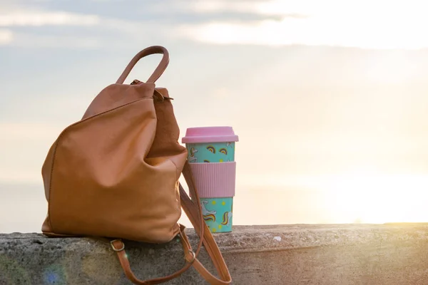 Bag and tumbler cup with coffee against blurred cityscape during sunrise. Girl traveler objects concept, copy space.