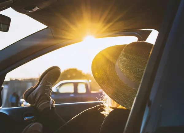 Blonde woman in hat resting in car at sunset time