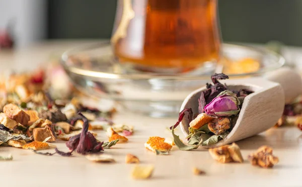 Mixed dry tea herbs and flowers in wooden spice shovel with unfocused tea in Turkish glass on background.