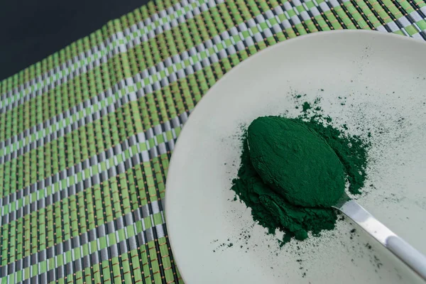 Spirulina powder on spoon on white plate with black background. Angled image. Natural organic spirulina supplement, superfood concept. — Stock Photo, Image