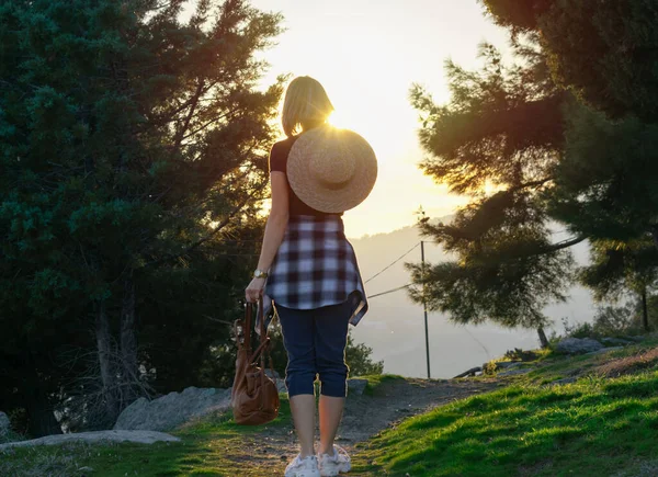 夕日の間に緑の公園を歩いてわらの帽子とバックパックを持つ若い女性。アクティブライフスタイルコンセプト. — ストック写真