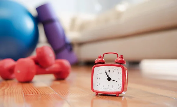 Alarm clock on the wooden floor with unfocus sports equipment and couch on background. Time for exercising concept