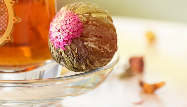 Sluiten bloeiende jasmijn groene thee bal met roze bloem op glazen schotel met turkse kopje thee. Wazige achtergrond, kopieer ruimte. Aziatisch thee genot concept. — Stockfoto
