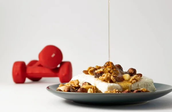 Soya cottage cheese with dry nuts and drop of honey falling on black plate. Unfocused red dumbbells on background. Protein nutrition vegan sports food.