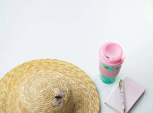 Straw hat with butterfly, pink tumbler cup and notebook with pen on white background with copy space on top. Girly summer objects concept.