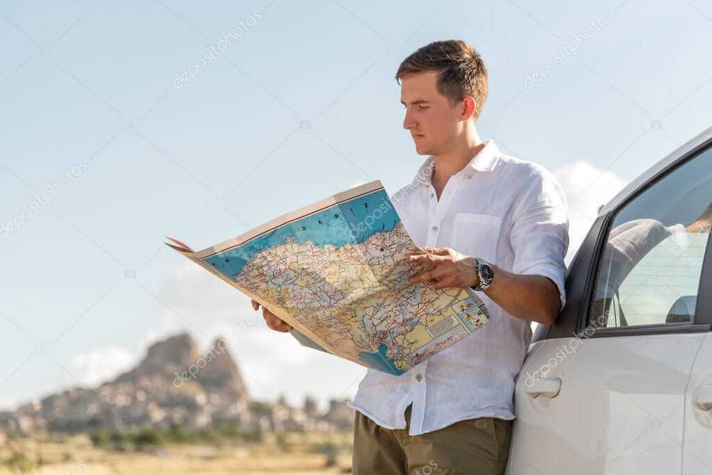 Caucasian young man standing byt the car looking at paper map, Uchisar city on backgorund. Lost traveler concept.
