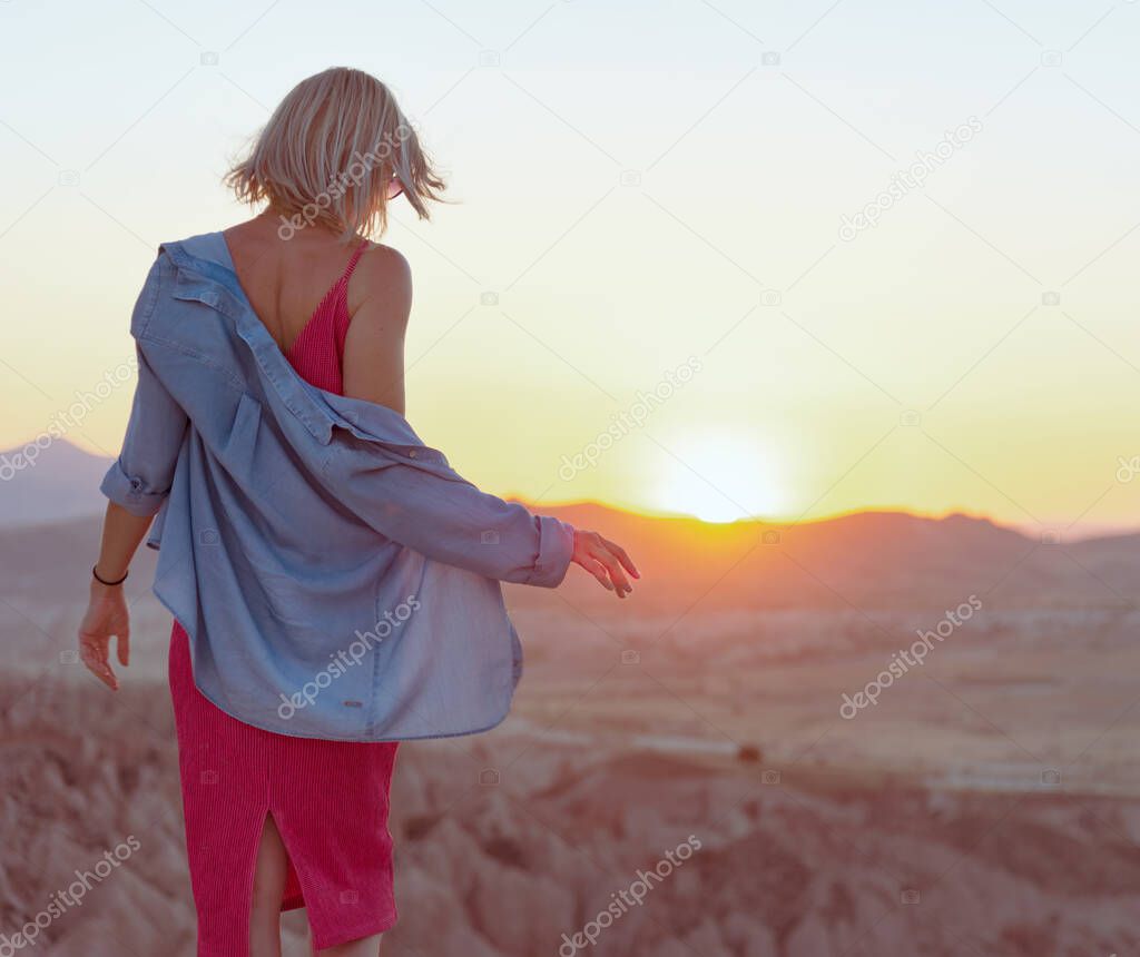 Back view of the young woman with blue shirt and pink dress standing at edge of the cliff and enjoying beautiful view of valley during sunset