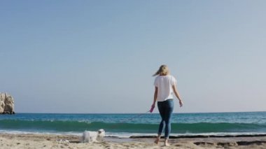 Güneşli bir günde beyaz Maltese köpeğiyle denize doğru yürüyen kadının yavaş çekimleri..