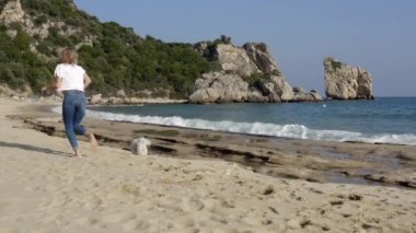 Güneşli bir günde sahilde beyaz Maltese köpeğiyle koşan gündelik elbiseli mutlu bir kadın. Yavaş çekim stok videosu.