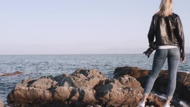 海によって岩の近くを歩く肩の上にDslrカメラとグランジスタイルの女性 — ストック動画