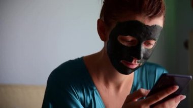 Elinde ipek bornoz ve spirulina maskeli genç bir kadının akıllı telefonuna bakarken çekilmiş görüntüleri..