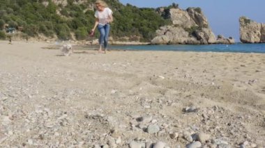 Güneşli bir günde sahilde beyaz Maltese köpeğiyle koşan gündelik elbiseli mutlu bir kadın. Yavaş çekim stok videosu.