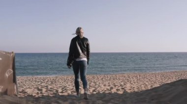 Grunge tarzı bir kadın, omzunda DSR kamerasıyla deniz kenarında kayaların yanında yürüyor..