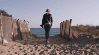 Grunge tarzı bir kadın, omzunda DSR kamerasıyla deniz kenarında kayaların yanında yürüyor..