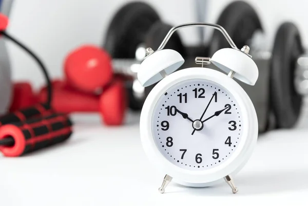 Yerde beyaz çalar saat ve arka planda odaklanmamış spor ekipmanları var. Egzersiz yapma zamanı.