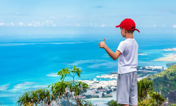 Kid with red baseball hat showing thumbs up gesture against scenic view of ocean bay from above.