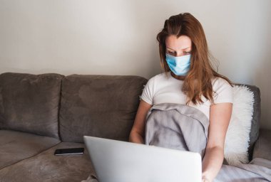 Tıbbi maske takan bir kadın dizüstü bilgisayarla koltukta oturuyor. Covid-19 salgını. Evde kal..