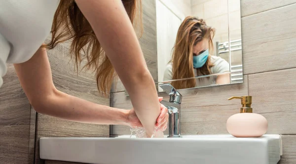 Woman with medical face mask washing hands with soap in the sink.
