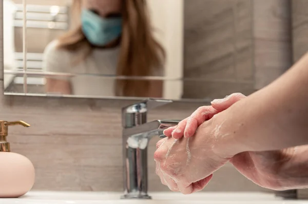 Woman with medical face mask washing hands with soap in the sink.