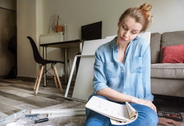 Konsantre genç bayan oturma odasında mobilyaları monte etme talimatlarını okuyor.