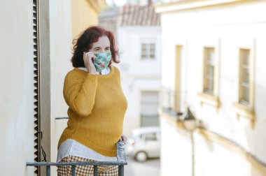 Koruyucu maskeli kadın balkonda cep telefonuyla konuşuyor.