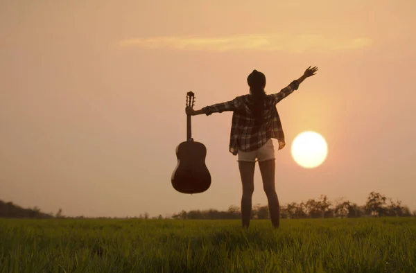シルエット女性ミュージシャン保持ギターと立っている緑の芝生の上で日没や日の出の背景とともにコピースペース — ストック写真