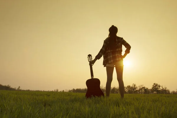 Silhouette女性ミュージシャン保持ギターと立って緑の芝生の上で日没や日の出の背景とともにコピーSpac — ストック写真