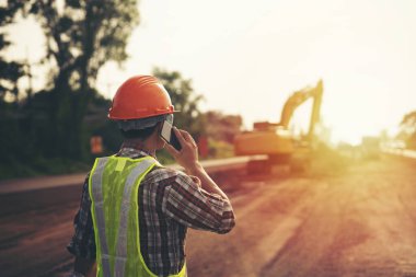 Mühendis sert bir cehennem taşı takıyor ve yol inşaatında makineli akıllı telefon kullanıyor.
