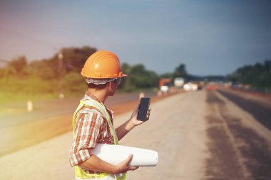 Mühendis sert bir cehennem taşı takıyor ve yol inşaatının planlarını makine ve güvenlik kavramlarıyla elinde tutuyor.