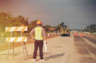 Mühendis sert bir cehennem taşı takıyor ve yol inşaatının planlarını makine ve güvenlik kavramlarıyla elinde tutuyor.