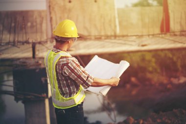 Mühendis sert bir cehennem taşı takıyor ve inşaat alanının planlarını tutuyor.