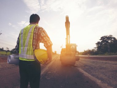 Mühendis sert bir cehennem taşı takıyor ve yol inşaatının planlarını makine ve güvenlik kavramlarıyla elinde tutuyor.