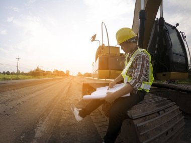Mühendis sert bir cehennem taşı takıyor ve yol inşaatının planlarını makine ve güvenlik kavramlarıyla elinde tutuyor.