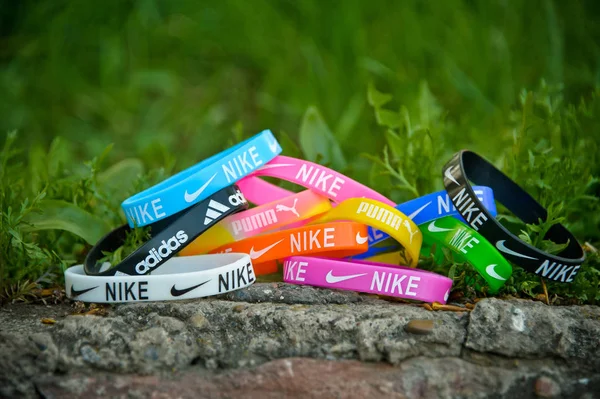 Färgglada elastiska gummiarmband, armband med etiketter av kända märken Nike, Adidas, Puma skott på klipporna och grönt gräs bakgrund. Regnbåge silikon runda armband bära närbild. — Stockfoto