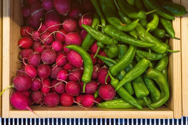 Green chili peppers and red radishes in wooden box on counter top of a store.