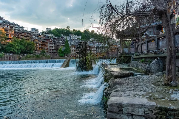 Çin 'in Hunan bölgesindeki eski Fenghuang Antik Kasabası' nda nehrin kenarındaki eski bir su değirmeninin önünde bulutlu bir gün batımı.