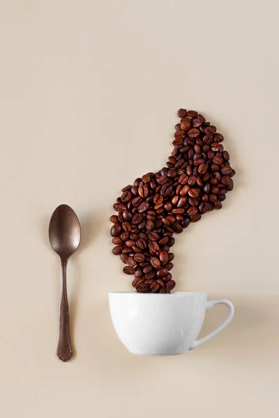 Творческий кофе натюрморт. Кофейные зерна выкладываются над чашкой в виде пара. Рядом лежит бронзовая или металлическая ложка, творческая концепция — стоковое фото