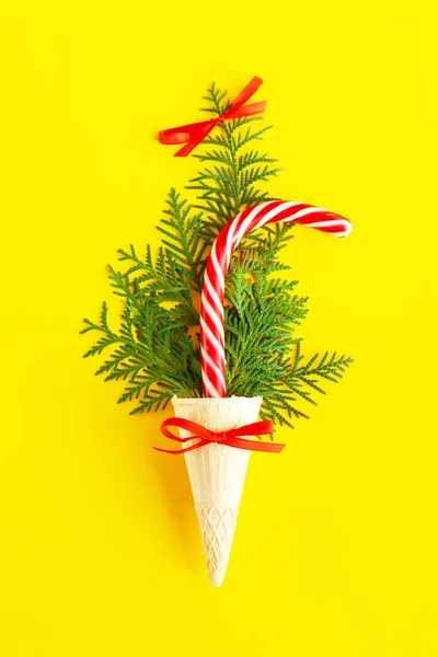 Striped red and white Christmas cane and thuja, fir or spruce branches in a waffle cone for ice cream on the yellow background. Creative, festive still life with food, flat lay