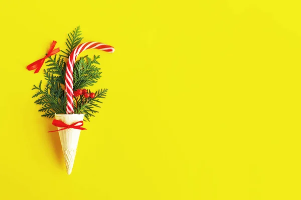 Striped red and white Christmas cane and thuja, fir or spruce branches in a waffle cone for ice cream on the yellow background. Creative, festive still life with food, flat lay, copy space