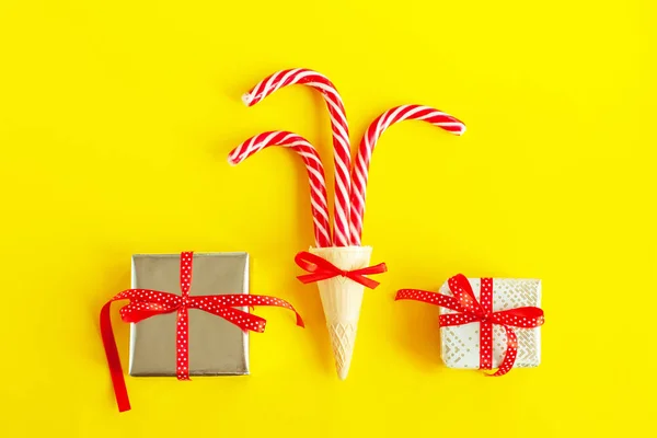 A waffle ice cream cone with three Christmas canes and wrapped gifts or boxes with red ribbons and bows on a bright, juicy yellow background. Creative, festive flat lay concept