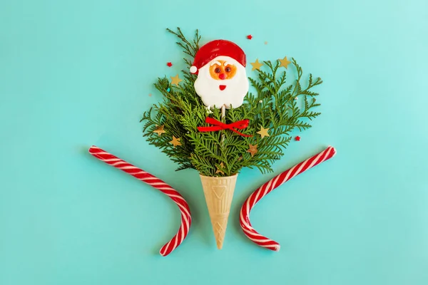 Waffle cone for ice cream is filled with Christmas tree branches, star-shaped confetti, a lollipop in the shape of Santa Claus, next to it are two Christmas canes. Funny flat lay on a light blue background