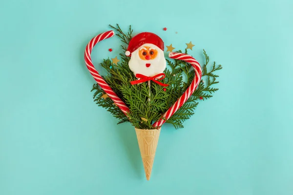 The waffle cone for ice cream is filled with Christmas tree branches, star-shaped confetti, a lollipop shaped like Santa Claus and Christmas canes. Holidays flat lay on a light blue paper background