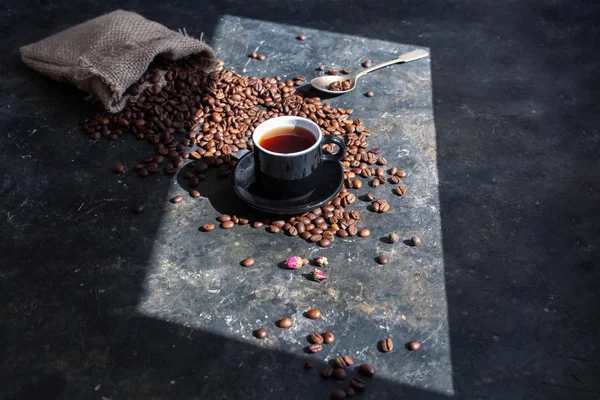 Beautiful still life with cup of morning coffee and ingredients for making coffee on a dark concrete surface. Coffee beans spill out of the bag in the background. Play of light and shadow, unusual lighting — 스톡 사진