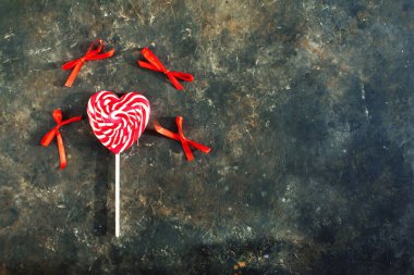 Tek bir beyaz-kırmızı çizgili lolipop. Koyu renkli beton arka planda kalp şeklinde, üst görünümde. Şekerlerin etrafında kelebekler gibi kırmızı fiyonklar var. Sevgililer Günü ya da aşk konsepti