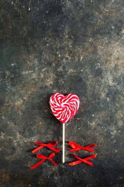 Beyaz-kırmızı çizgili lolipop. Koyu renkli beton arka planda kalp şeklinde. Şekerlerin yanında dört kırmızı fiyonk var, üst manzara. Sevgililer Günü konsepti, boşluğu kopyala
