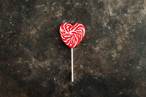 Single white-red striped lollipop in the shape of a heart on a dark creative concrete background. Top view. Valentines day or love concept. Minimalistic greeting card or advertisement design