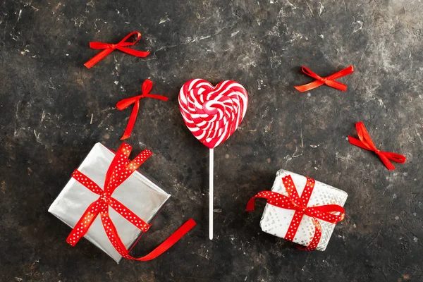 White-red striped lollipop in the form of a heart and festive wrapped gifts on a dark aged background, flat lay. Around the candy are red bows. Valentines day concept. Love for shopping, close up