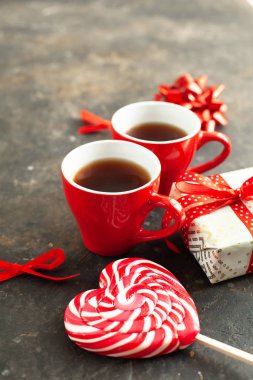 İki fincan sıcak espresso, kahve ya da tişört, güzel paketlenmiş hediye, kalp şeklinde beyaz-kırmızı lolipop ve desenli arka planda dekoratif fiyonklar. Sevgililer Günü romantik randevu konsepti, yakın çekim.