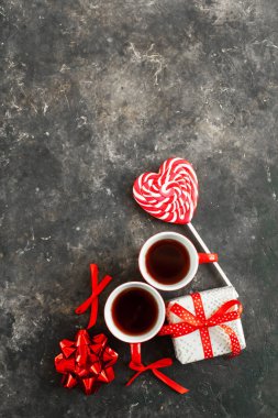 İki fincan espresso ya da kahve, paketlenmiş hediye, kalp şeklinde beyaz-kırmızı lolipop ve fotoğrafın altına yerleştirilmiş dekoratif fiyonk yaratıcı beton arka planda. Boşluğu kopyala